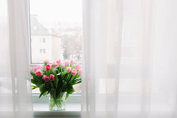 Bouquet of pink tulips in transparent vase on windowsill, against background of window. Flowers in interior.