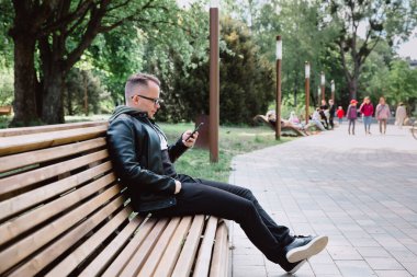 Siyah kot pantolon ve deri ceketli genç beyaz adam şehir parkında bankta oturuyor, akıllı telefonuyla internet üzerinden haberleri okuyor. Kentsel bir çevrede çalışmaktan dinlenme.