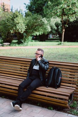 Siyah kot pantolonlu, deri ceketli, sırt çantalı, parktaki bankta oturan ve akıllı telefondan konuşan beyaz bir genç. Kentsel bir ortamda dinlenmek, şehir hayatı.