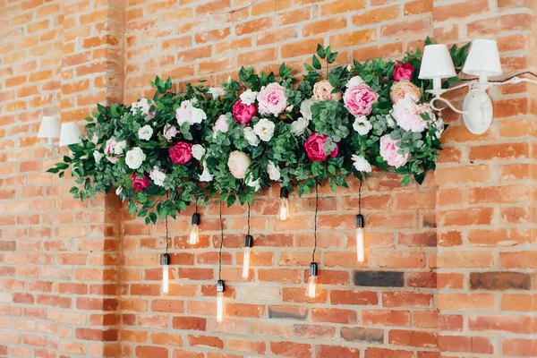 Taze çiçeklerin ve parlayan ampullerin çiçekli kompozisyonu tuğla duvarı süslüyor. Kırsal kutlama dekorasyonu. Seçici odak.