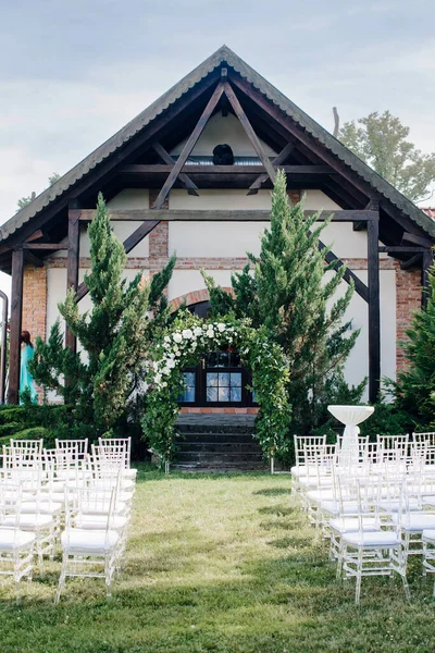 Düğün dekoru, doğada bir düğün töreni için bir yer, bahçede. Taze çiçeklerden yapılmış düğün kemeri, çiçekçi ve misafirler için şeffaf sandalyeler. Dikey fotoğrafçılık.