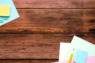 Ahşap arka plandaki ofis eşyalarından yapılmış çerçeve: renkli kağıt, kalem, ataç. Düzen, metin için boş alan.