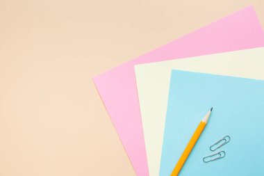 Ofis masasında bir takım malzemeler, renkli kağıtlar, kalem, bej arka planda ataç. Üst görünüm, metin için boş boşluk.