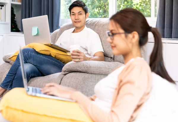 Asian working man use computer notebook laptop work from home in living room with social distance to his couple in background while city lockdown. Work at home concept.