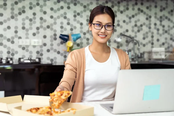 Asian woman work from home in the kitchen and eating delivery pizza take away food and take out coffee while city lockdown from coronavirus covid-19 pandemic. New Normal lifestyle working from home.