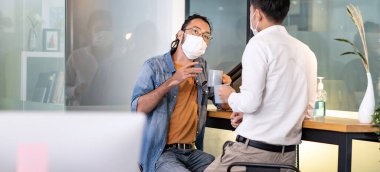 Panoramik iki ofis çalışanı konuşurken, kahve molası, sosyal mesafe muayenehanesi ile yeni normalleşecek. Yüz maskesi takıyorlar COVID-19 koronavirüsünün riskini azaltmak için..