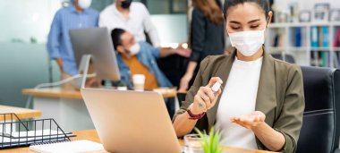 Panoramik Asyalı ofis çalışanı iş kadını yüz maskesi takıyor. Yeni normal ofis ortamında hijyen için alkol spreyi kullanıyor. Coronavirüs COVID 19 'un yayılmasını engelliyor..