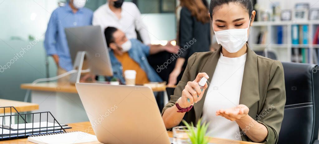 Panoramic Asian office employee businesswoman wear face mask use alcohol spray hand sanitizer for hygiene in new normal office with social distance practice prevent coronavirus COVID-19 spreading.