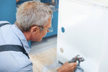 Bir çalışan metal ürünleri boyuyor. Yaşlı bir adam elinde kompresör sprey tabancası tutuyor ve kabineyi beyaza boyuyor.