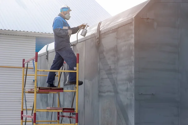 Industrial work. Priming of metal products from the compressor gun. A worker in overalls and a respirator stands on the stairs and paints the body of a truck trailer or a metal car. Work at height