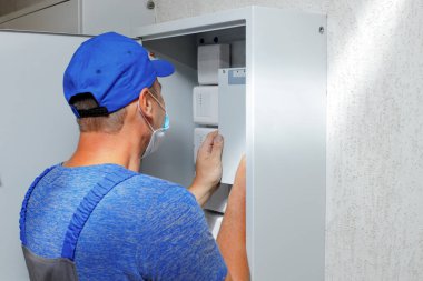 Tulumlu ve beyzbol şapkalı bir elektrikçi, sensörleri ve ekipmanları olan bir kabineyi inceliyor. Elektrik ve yangın alarmlarının teşhisi ve bakımı. Düzen ya da maket.