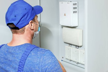 Tulumlu ve beyzbol şapkalı bir elektrikçi, sensörleri ve ekipmanları olan bir kabineyi inceliyor. Elektrik ve yangın alarmlarının teşhisi ve bakımı. Düzen ya da maket.