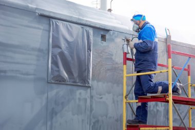 Endüstriyel işler. Kompresör silahından çıkan metal ürünlerin hazırlanması. Tulumlu ve solunum cihazlı bir işçi merdivenlerde durur ve bir kamyon römorku ya da metal bir arabanın vücudunu boyar. Yükseklikte çalış