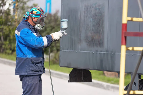 Industrial work. Priming of metal products from the compressor gun. A worker in overalls and a protective mask paints the body of a truck trailer or a metal car. Not staged photo