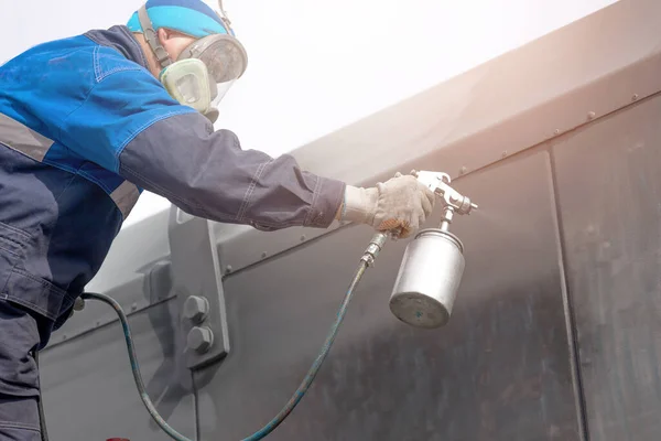 Industrial work. Priming of metal products from the compressor gun. A worker in overalls and a protective mask paints the body of a truck trailer or a metal car. Not staged photo