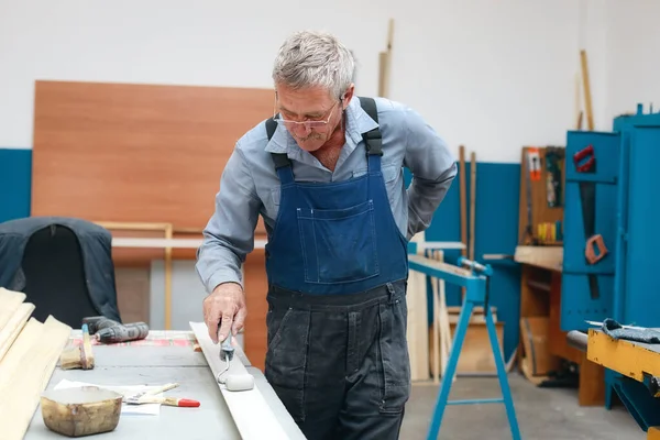 Tulum ve gözlüklü yaşlı bir marangoz dükkanındaki tezgahta bir silindir ile ahşap bir tahta boyuyor.. — Stok fotoğraf