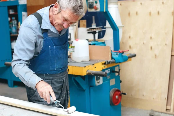 Un anciano ebanista en mono y gafas pinta una tabla de madera con un rodillo en un banco de trabajo en una carpintería. — Foto de Stock