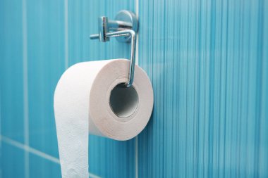 A roll of toilet paper hangs on a metal holder against a blue tile wall.  clipart