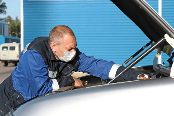 Білий чоловік у медичній масці на обличчі ремонтує машину на вулиці у відкритому космосі . — стокове фото