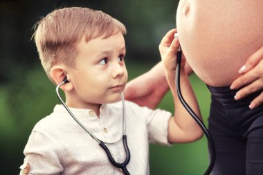 Küçük bir çocuk, hamile bir annenin steteskopuyla midesini dinler..