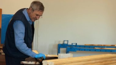 An elderly carpenter with glasses varnishes wooden boards in a carpenters shop.
