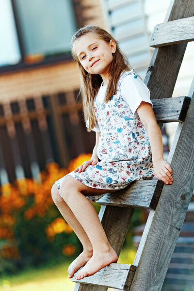 A country girl in a summer dress sits on a wooden staircase on a summer day and looks into the camera.