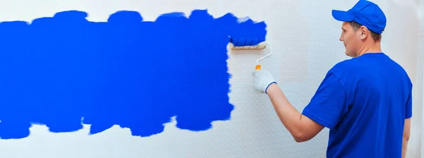배너 건설 및 수리. 제복을 입은 남자가 페인트 롤러로 방 안의 벽을 파란색으로 물들이고 있습니다.. — 스톡 사진