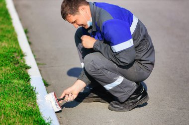 Tulumlu bir işçi yaz günü kaldırımı beyaz boyayla boyar. Şehir hizmeti, bölgelerin iyileştirilmesi.