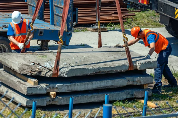 Dwóch procy rozładowuje betonowe płyty na ulicy w letni dzień. Pracownicy w kaskach budowlanych i kamizelkach ładunkowych procy. — Zdjęcie stockowe