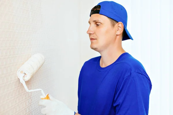 Ένας ζωγράφος με καπέλο και γάντια βάφει τον τοίχο με ρολό μπογιάς. Προσωπογραφία ενός νέου εργάτη με στολή. — Φωτογραφία Αρχείου