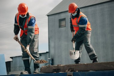 İki işçi iş kıyafetleri ve inşaat alanında küreklerle tıbbi maske takıyor. Ağır iş gücü..