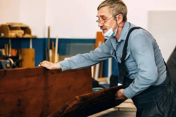 A gray-haired carpenter with glasses works with wood on a workbench in a carpentry shop.