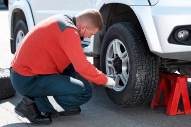 Tıbbi maskeli bir araba tamircisi güneşli bir günde caddedeki bir arabanın tekerleğini değiştirir. Yardım yolda.