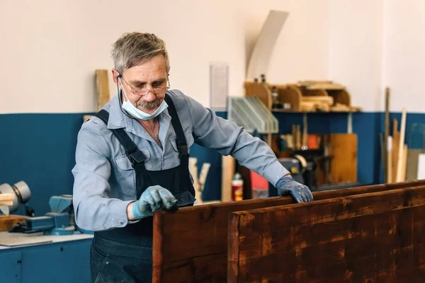 Пожилой человек рисует деревянные доски с темным лаком в столярной мастерской. Пенсионер в медицинской маске — стоковое фото