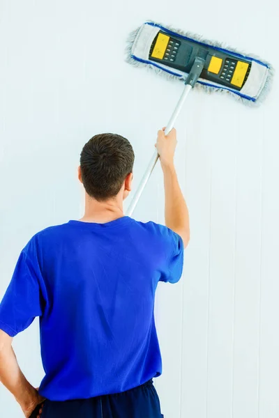 Ένας νεαρός λευκός με μπλε μπλουζάκι πλένει τον λευκό τοίχο του δωματίου με μια σφουγγαρίστρα.. — Φωτογραφία Αρχείου