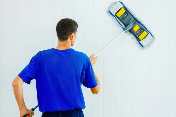 Egy fiatal fehér férfi kék pólóban mossa a szoba fehér falát egy felmosóval.. — Stock Fotó