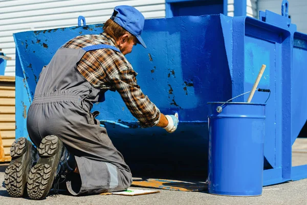 İş tulumu giymiş yaşlı bir adam sokağa boya fırçasıyla çöp tenekesi ya da konteynır boyuyor.. — Stok fotoğraf