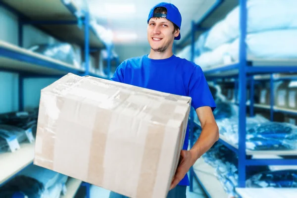 Een jonge positieve magazijnmedewerker houdt een kartonnen doos in zijn handen en glimlacht. — Stockfoto