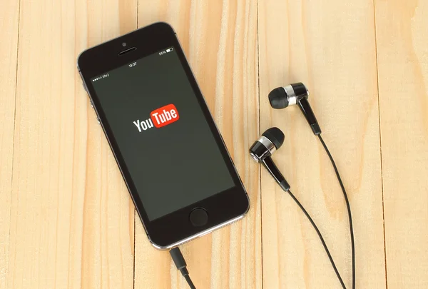 Teléfono inteligente con logotipo de YouTube en su pantalla y auriculares sobre fondo de madera — Foto de Stock