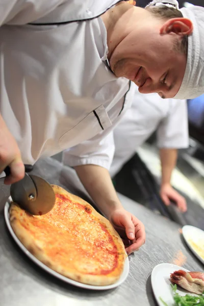 Tangan dekat koki tukang roti dengan seragam putih memotong pizza di dapur — Stok Foto
