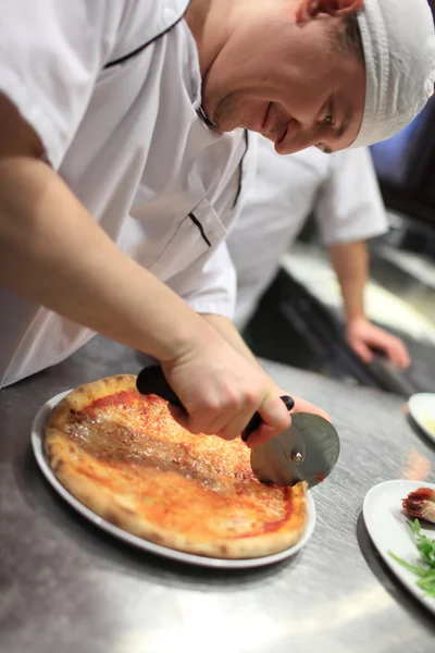 Zbliżenie ręka szefa kuchni piekarz w białym mundurze cięcia pizzy w kuchni — Zdjęcie stockowe