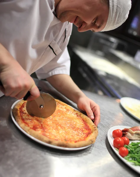 Mão fechada de padeiro chef em uniforme branco cortando pizza na cozinha — Fotografia de Stock