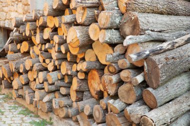 Büyük, düzgünce istiflenmiş taze kesilmiş odun yığınları dışarıda taş bir duvarın yanında. Doğal ahşap dokusu ve toprak renkleri, kışın ısıtma ve sürdürülebilir enerjiyi sembolize eden kırsal bir arka plan sağlar..