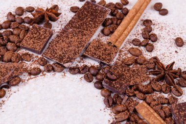 Beyaz bir masanın üzerinde kavrulmuş kahve çekirdekleri, baharatlar, tarçın çubukları ve kakao tozuyla kaplı anason yıldızlarıyla çikolata parçaları. Köşedeki boşluğu daire şeklinde kopyala.