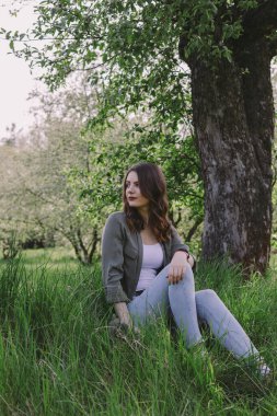 Çekici esmer kadın bahçedeki ağacın altındaki çimlerin üzerinde oturuyor..