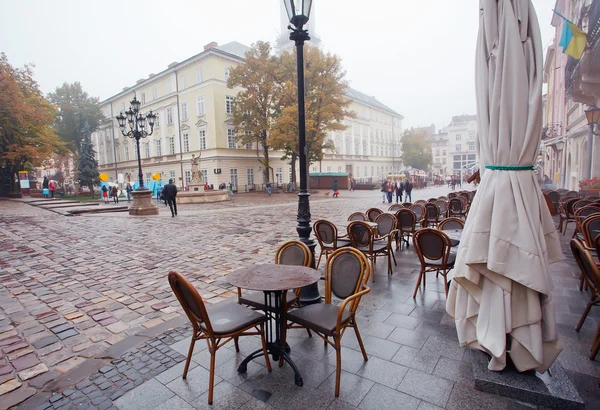 People walk under rain at Market Square with cobbled streets and empty outdoor cafe — Stock Photo, Image