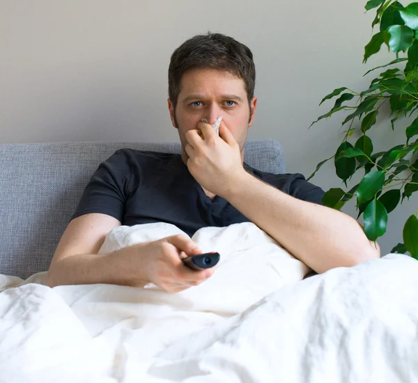 Homem doente soprando o nariz enquanto assiste TV em casa . — Fotografia de Stock