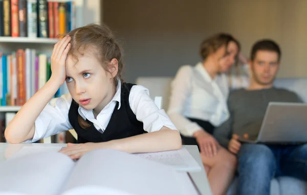 Müdes kleines Mädchen bei den Hausaufgaben, während ihre Eltern auf dem Sofa liegen. — Stockfoto