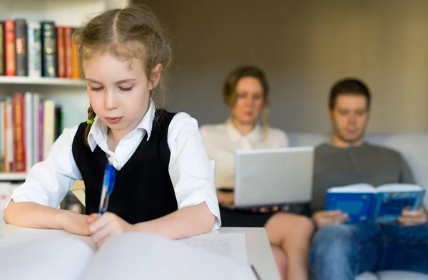 Petite fille faisant ses devoirs, tandis que ses parents se reposent sur le canapé . — Photo