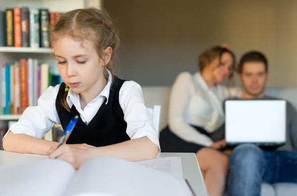 Petite fille faisant ses devoirs, tandis que ses parents se reposent sur le canapé . — Photo